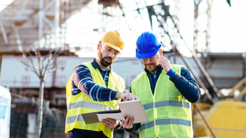 Two construction workers in safety vests and helmets review plans at a construction site.