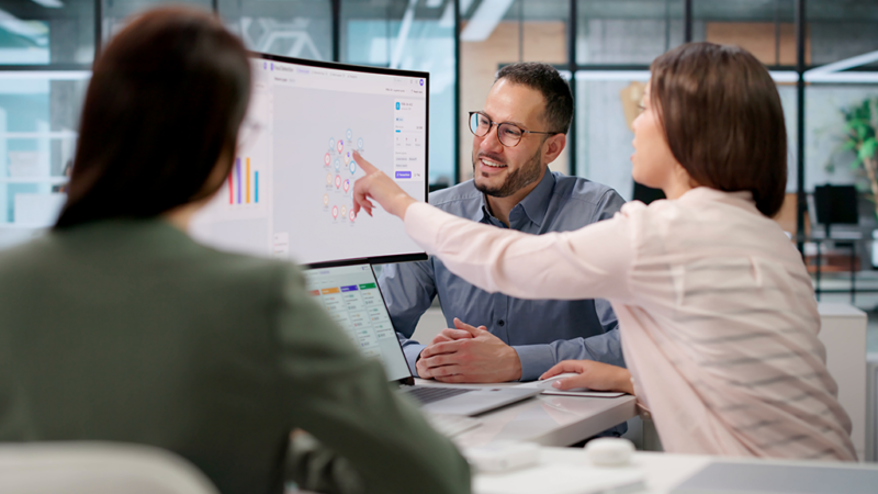Three colleagues review data on a large monitor in a modern office as a woman points at the screen and a bearded man in glasses smiles.